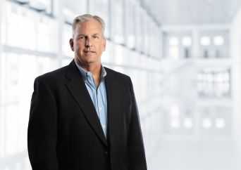 A middle-aged man wearing a black blazer and light blue shirt stands in a bright, modern, white hallway with large windows and soft natural light.