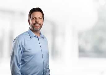 A smiling man with short dark hair and a beard, wearing a light blue button-up shirt, stands in front of a bright, blurred background.