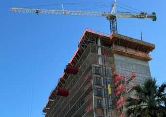A tall building under construction with scaffolding and a crane overhead, set against a clear blue sky. Some red safety barriers and palm tree leaves are visible in the foreground.