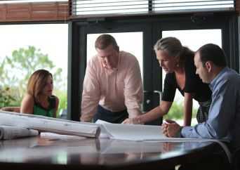 Four people gather around a table reviewing large architectural blueprints, engaged in discussion in a well-lit office with large windows in the background.