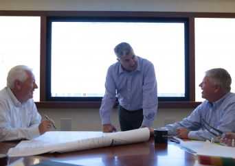 Three men in business attire are having a discussion around a conference table with documents and blueprints; one man stands while the others sit, all in front of a large bright window.