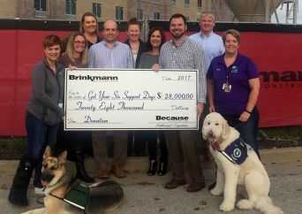 A group of people poses outdoors, holding a large donation check for $28,000 made out to “Got You Six Support Dogs.” Two support dogs wearing vests sit at the front of the group.