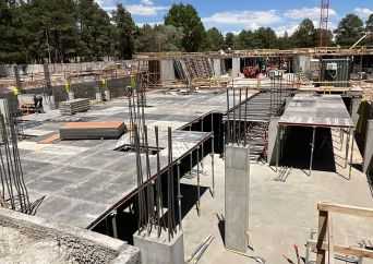 Construction site with concrete slabs, steel reinforcement bars, and partially built walls; trees and blue sky are visible in the background.