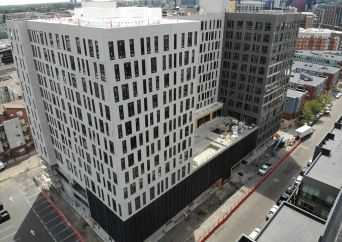Aerial view of a modern multi-story building complex with white and dark facades, featuring many rectangular windows, under construction along a city street.