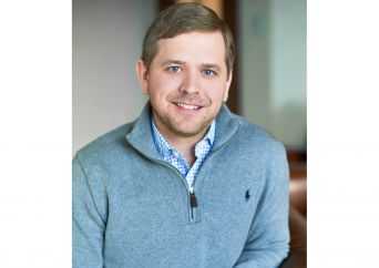A man with short light brown hair and a beard, wearing a light gray quarter-zip sweater over a blue patterned shirt, smiling and sitting indoors.