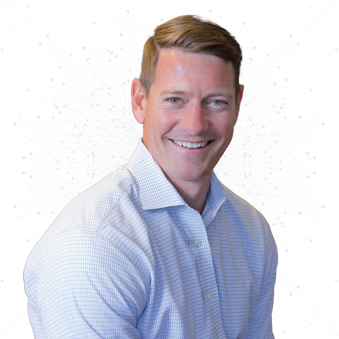 A man with short light brown hair, wearing a light blue checkered dress shirt, smiles at the camera against a white background with faint circular patterns.