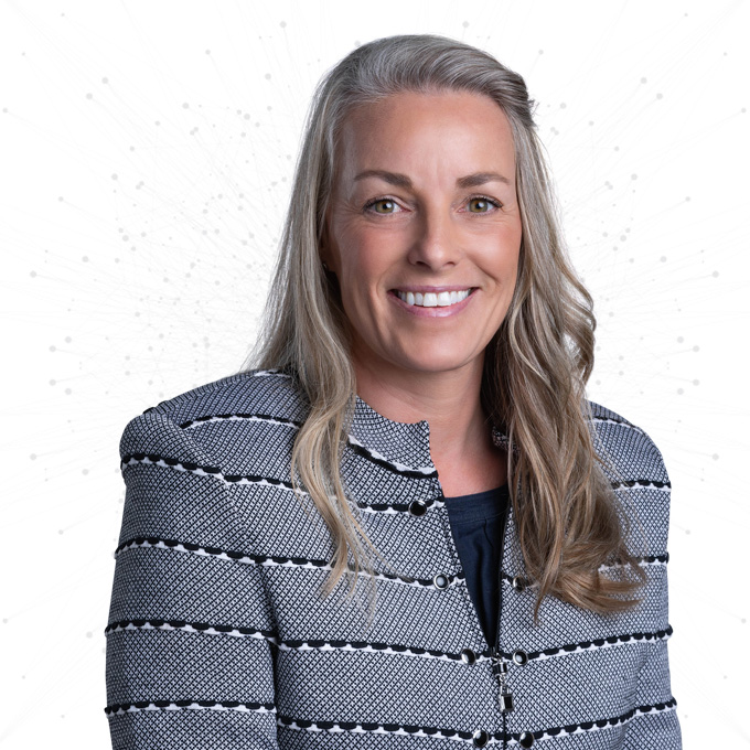 A woman with long, wavy blonde hair wearing a patterned blazer and a dark top smiles at the camera against a white background with subtle abstract dots.