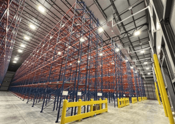 A large, empty warehouse with tall blue and red metal storage racks under bright overhead lights. Yellow safety barriers are positioned at the end of the racks on a clean concrete floor.