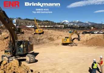 Construction site with three excavators digging in a large dirt pit. Three workers in safety gear stand nearby. Snow-capped mountains and vehicles are visible in the background. Logos and rankings for ENR and Brinkmann Constructors appear on the image.