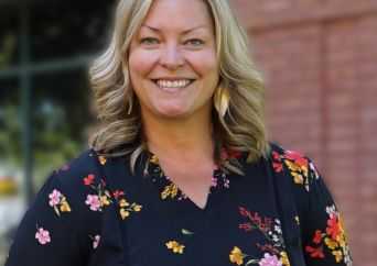 A woman with blonde hair wearing a black floral top smiles outdoors in front of a brick wall and a window.