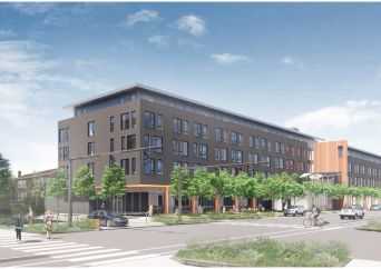 A modern four-story apartment building with large windows and a brown exterior stands at a street corner, surrounded by trees, pedestrians, cyclists, and a few cars under a partly cloudy sky.