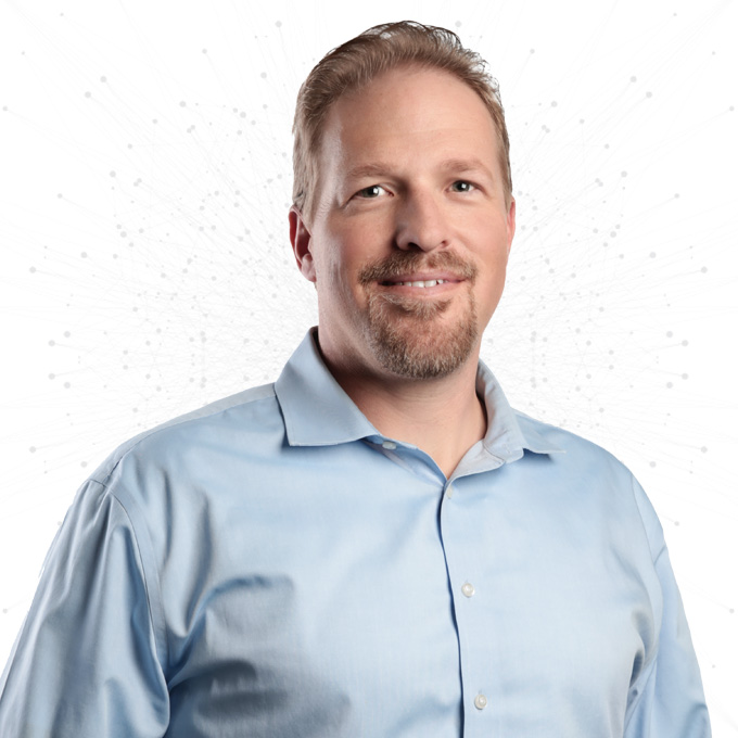 A smiling man with short light brown hair and a light beard, wearing a light blue button-up shirt, stands in front of a white background with subtle dotted patterns.