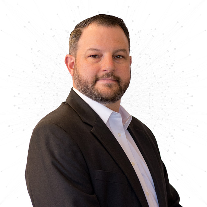 A man with short dark hair and a beard, dressed in a white shirt and dark blazer, poses confidently against a white background with subtle gray dot and line patterns.
