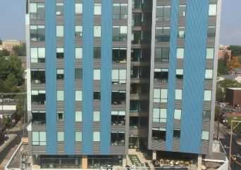 A modern mid-rise building with blue and gray panels, large windows, and an outdoor seating area at the ground level. Surrounding trees and other buildings are visible in the background.