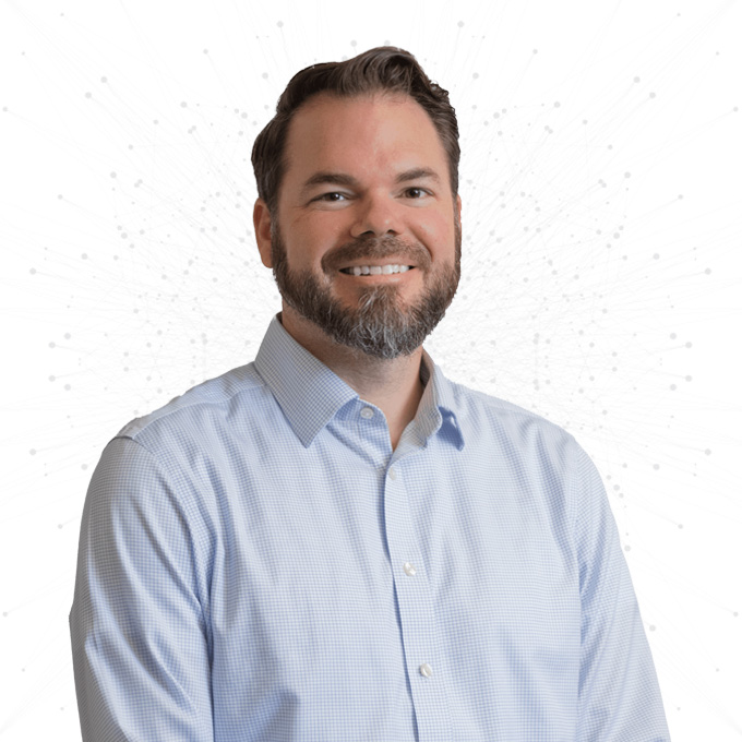 A man with short brown hair and a beard, wearing a light blue button-up shirt, smiles while standing against a white background with a faint network pattern.
