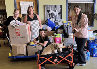 Four smiling women pose with a cart full of donated supplies; one woman is holding a wagon with a tan dog inside. Boxes, bags, and pet food are visible around them indoors.