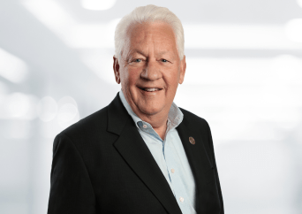 Smiling older man with short white hair wearing a dark suit jacket and light blue collared shirt, standing in front of a bright, blurred background.
