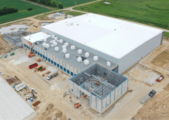 Aerial view of a large industrial building under construction, surrounded by dirt and construction vehicles, with green fields in the background. The structure has a white roof and sections with visible steel framing.