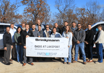 A group of people stand outdoors in front of trees, holding a banner that reads “Brinkmann, OASIS AT LAKEPORT, PARTNERSHIP.” Two vehicles are visible in the background, and everyone is smiling at the camera.