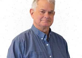 Older man with short gray hair wearing a blue checkered button-up shirt, standing and facing the camera with a neutral expression against a plain white background.