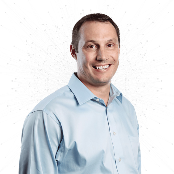 A smiling man wearing a light blue button-up shirt stands against a white background with a subtle network pattern.