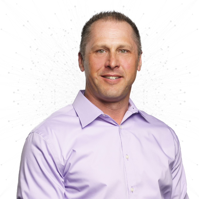 A man with short brown hair wearing a light purple button-up shirt stands in front of a white background with faint abstract dot patterns. He is smiling slightly and facing the camera.