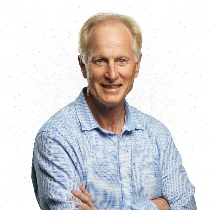 An older man with light hair and a friendly smile stands with arms crossed, wearing a light blue checkered shirt. The background is white with a faint abstract pattern of dots and lines.