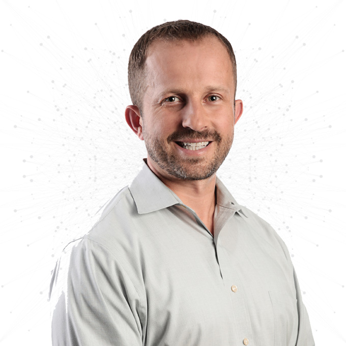 A man with short hair and a trimmed beard is smiling, wearing a light gray button-up shirt, standing in front of a white background with a subtle dotted pattern.
