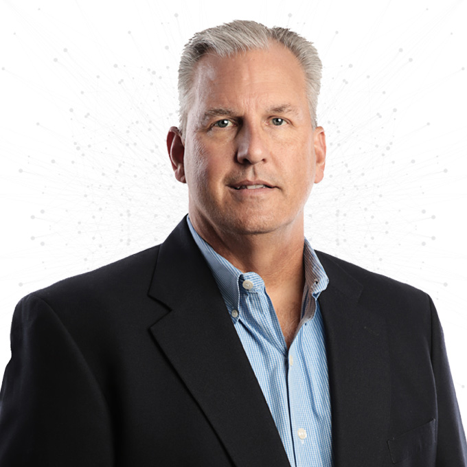 A middle-aged man with short gray hair, wearing a dark blazer over a light blue collared shirt, stands in front of a white background with a subtle network pattern.