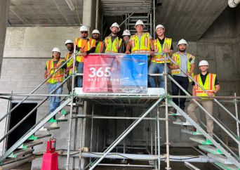A group of construction workers wearing safety vests and hard hats stand on scaffolding stairs around a sign that reads 365 Safe Days. They are posing for a photo at a construction site with concrete walls.