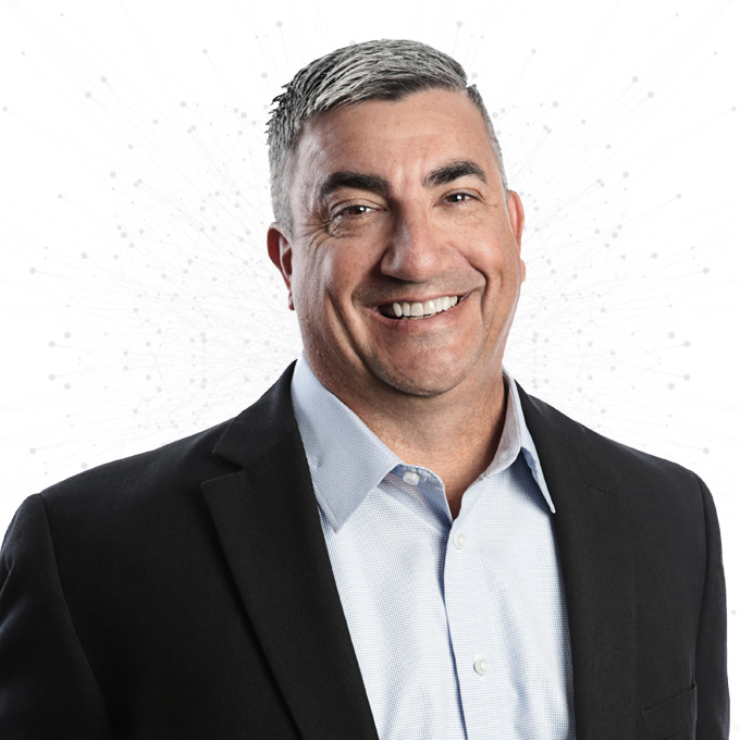 A smiling man with short gray hair wearing a dark blazer and light blue dress shirt, standing against a white background with a subtle network pattern.