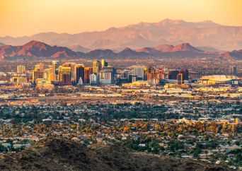 A wide view of a city skyline with tall buildings surrounded by desert and mountains in the background, all illuminated by golden sunlight.