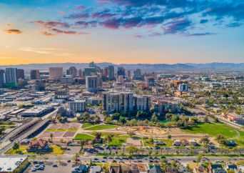 Aerial view of a cityscape with tall buildings, green parks, and roads under a colorful sunset sky with scattered clouds.