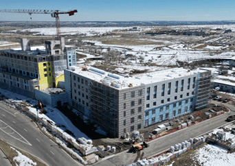 Aerial view of a multi-story building under construction with scaffolding and a tall crane, surrounded by snow-covered ground and roads, with distant fields and buildings in the background.