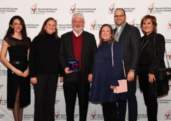 Six adults pose and smile together in front of a Ronald McDonald House Charities step-and-repeat backdrop; one person in the center holds a blue award plaque with a red heart.