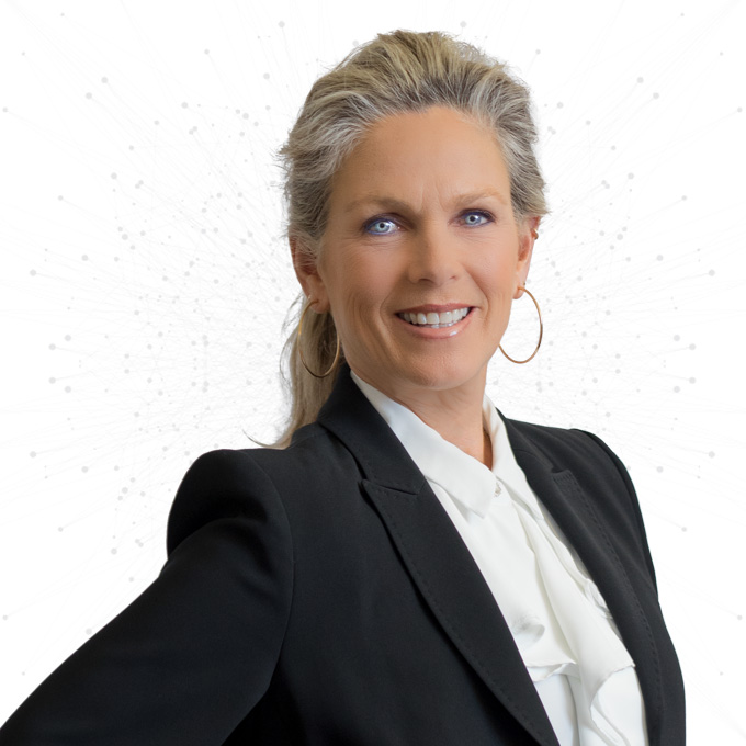 A woman with long, grayish-blonde hair in a ponytail, wearing a black blazer and white blouse, smiling confidently at the camera with a white background and subtle dotted design behind her.