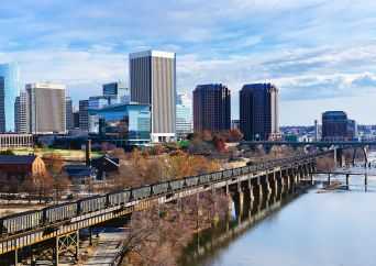 A city skyline with modern high-rise buildings, a wooden walkway bridge, and a river in the foreground under a partly cloudy blue sky.
