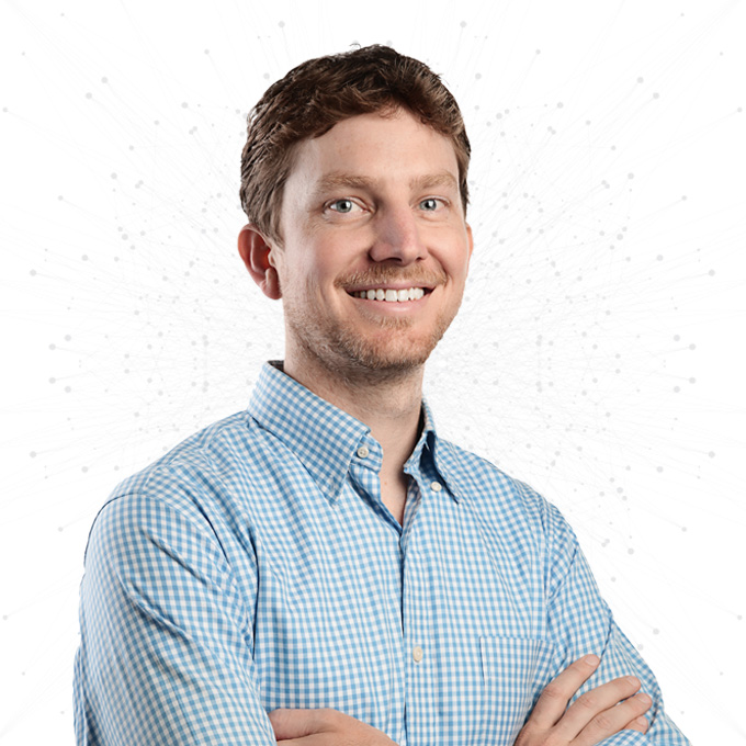A man with short, curly brown hair and a beard smiles with arms crossed, wearing a light blue checkered shirt, standing against a white background with a subtle dotted pattern.