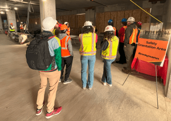 A group of people in safety vests and hard hats stand in a construction area, gathered near a sign that reads Safety Orientation Area. Some are listening to a speaker, while others stand nearby with backpacks.