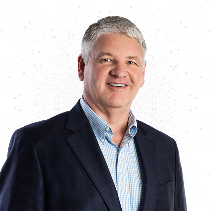 A middle-aged man with short gray hair wearing a blue checked blazer and light blue button-up shirt, smiling at the camera against a white background with a subtle dotted pattern.