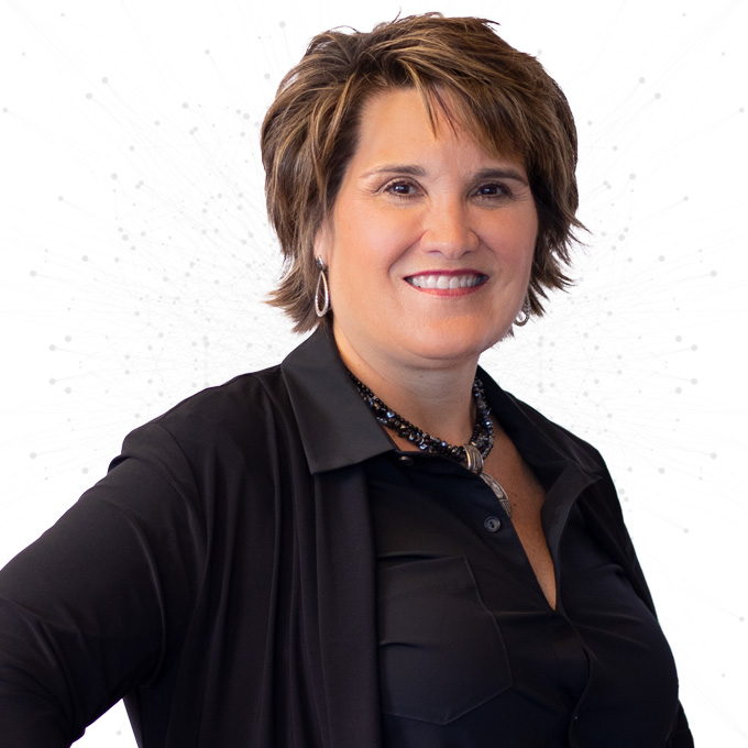 A woman with short brown hair smiles confidently. She is wearing a black button-up shirt, a black cardigan, a beaded necklace, and hoop earrings, standing against a light background with a subtle dotted pattern.
