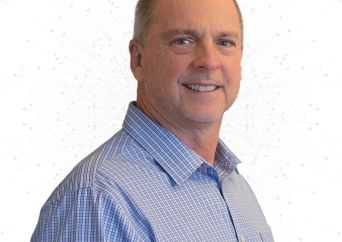 A middle-aged man with short light hair, wearing a blue checked button-up shirt, smiling and standing against a plain white background.