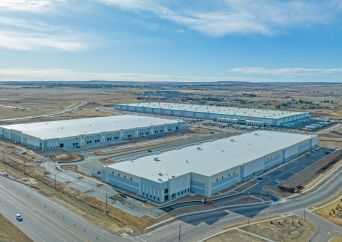 Aerial view of two large warehouse buildings surrounded by roads and parking lots in a mostly barren landscape under a partly cloudy sky.