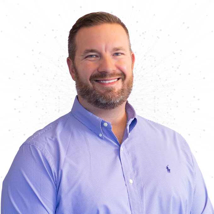 A smiling man with short brown hair and a beard, wearing a light purple button-up shirt, stands against a white background with a subtle abstract pattern.