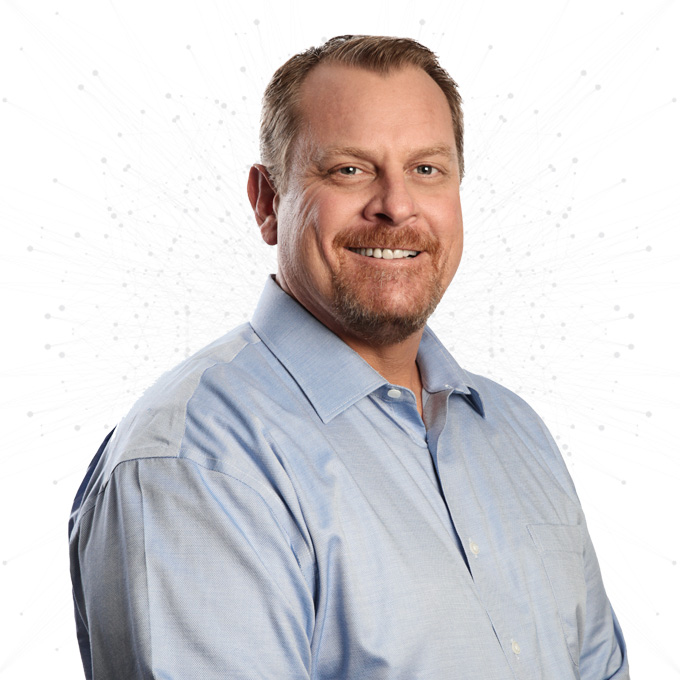 A smiling man with short light brown hair and a goatee, wearing a light blue button-up shirt, poses in front of a white background with a faint, abstract dotted pattern.