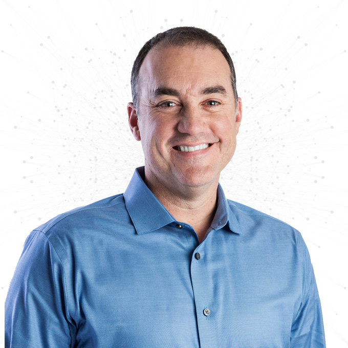 A man with short dark hair, wearing a blue button-down shirt, smiling at the camera. The background is white with a faint network-like design.