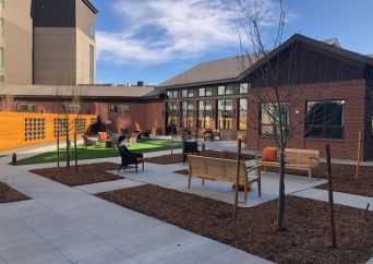 A modern outdoor courtyard with benches, chairs, and small trees, surrounded by buildings with large windows under a blue sky with scattered clouds.