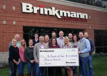 A group of people stands outside a brick building with a Brinkmann Constructors sign, holding a large check for $31,000 made out to Boys and Girls Club. Everyone is smiling at the camera.