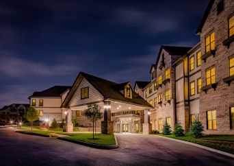 A large, well-lit building with multiple windows and peaked roofs is shown at night. Warm lights from inside illuminate the entrance, and neatly trimmed trees and grass line the curved driveway.