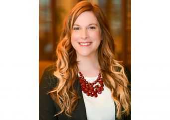 A woman with long, wavy light brown hair wearing a black blazer, white top, and a chunky red necklace, smiling at the camera with a blurred indoor background.